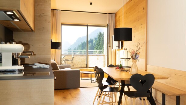 Una cocina moderna con cafetera, un comedor con mesa y sillas de madera, y un ventanal con vistas a montañas.