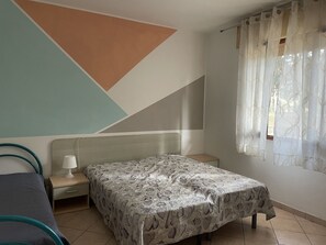 Une chambre avec un lit, une table de chevet, une lampe et une fenêtre ornée de rideaux légers.