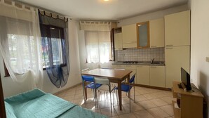 A small, well-lit room with a bed, a dining table with chairs, a kitchenette, and a TV on a stand. - Solaris by Interhome (Rosolina Mare)