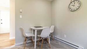 A room with a white table and two white chairs, a round wall clock, and a door in the background.