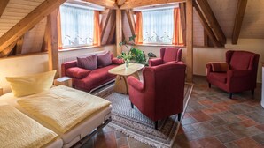 A cozy attic bedroom with a sloped ceiling, a red sofa, two red armchairs, a small table, and a bed with a beige blanket. - Eindverdieping DG (Sebnitz)