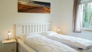 A white bed with a headboard, two bedside tables with lamps, a painting on the wall, and a window with curtains. - Beachkeeper Comfortable Holiday Residence (Zingst)