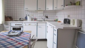 Een kleine keuken met witte kastjes, een fornuis, een spoelbak en een magnetron. Er staat een tafel met een geruit tafelkleed en een stoel.