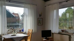 Een kamer met een eettafel, stoelen, een tv op een dressoir en grote ramen voorzien van gordijnen.
