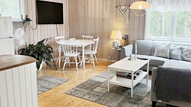 A room with a wooden floor, a white dining table with chairs, a grey sofa, a flat-screen TV mounted on the wall, and a window with curtains. - 5 Person Holiday Home in Djurhamn (Djurhamn)