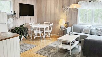 A room with a wooden floor, a white dining table with chairs, a grey sofa, a flat-screen TV mounted on the wall, and a window with curtains.