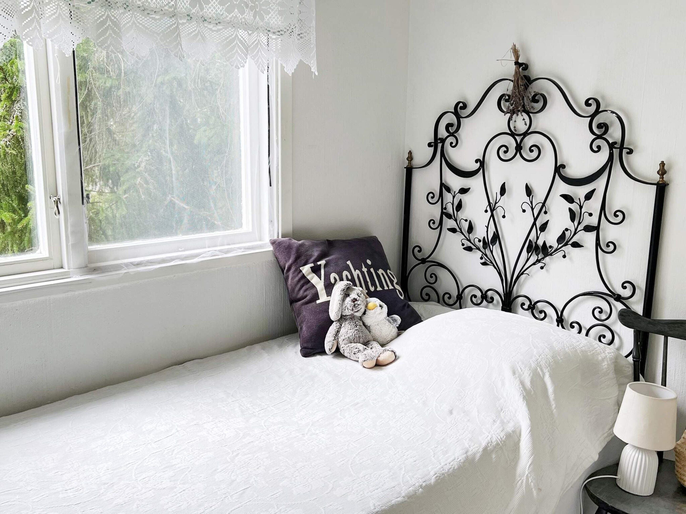 A bedroom with a single bed, a window with lace curtains, a decorative metal headboard, and a bedside lamp.