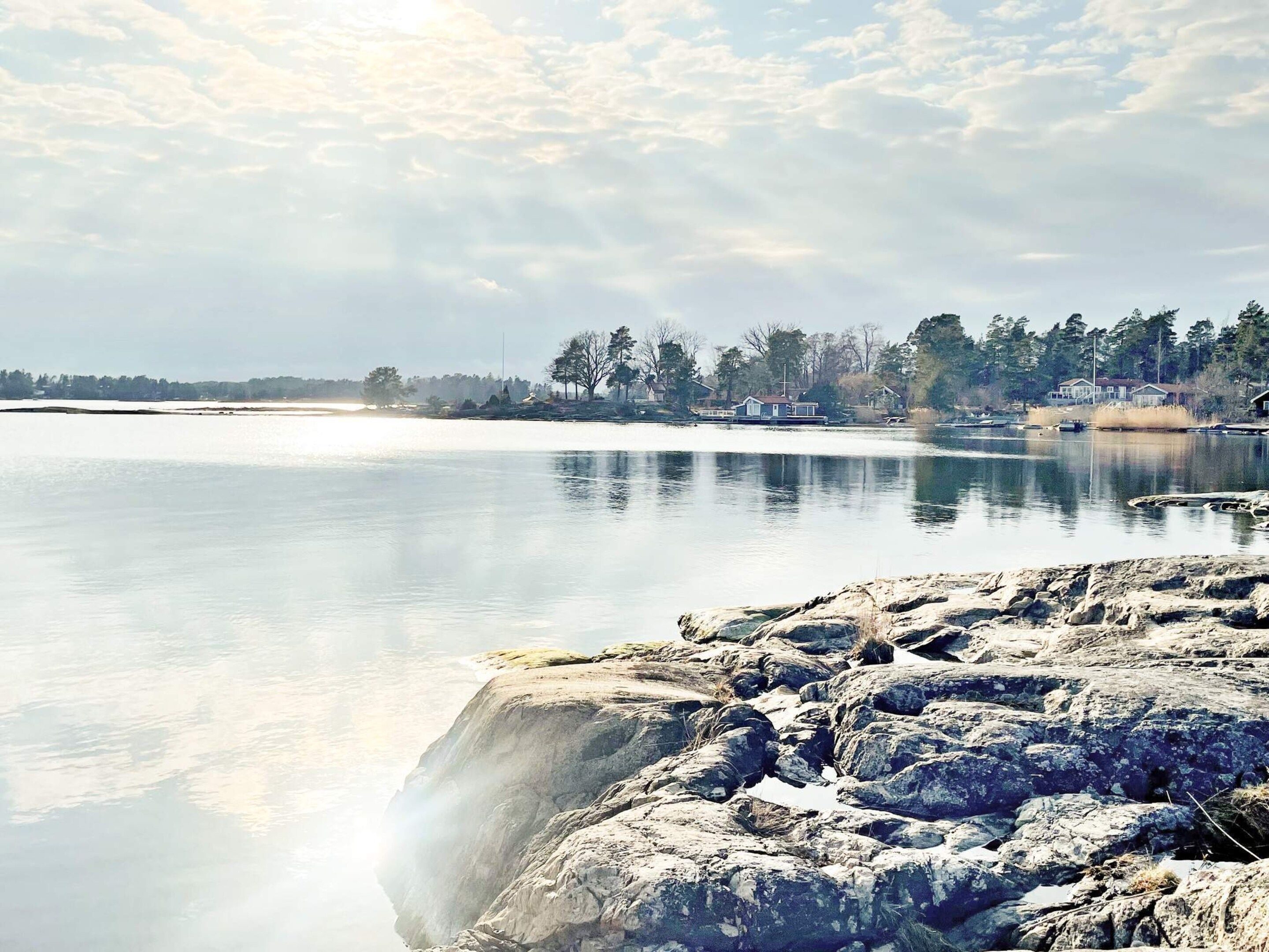 A calm lake with a rocky shoreline and a row of houses in the distance.