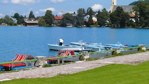 A lakeside view with boats docked along the shore, a church steeple in the background, and a stone pathway leading to the water. - Alpengluck am Wendelstein (Bayrischzell)