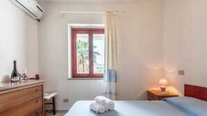Ett hotellrum med en säng, ett nattduksbord med en lampa, en byrå, ett fönster med gardiner och utsikt över palmer utanför.