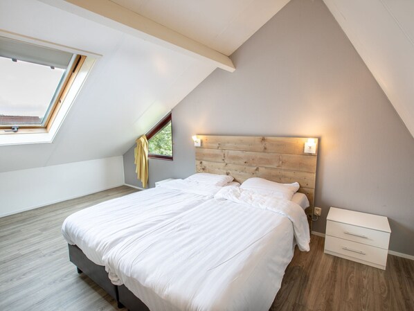 Quarto com teto inclinado, claraboia, cama com roupa de cama branca, cabeceira de madeira e mesa de cabeceira com abajur.