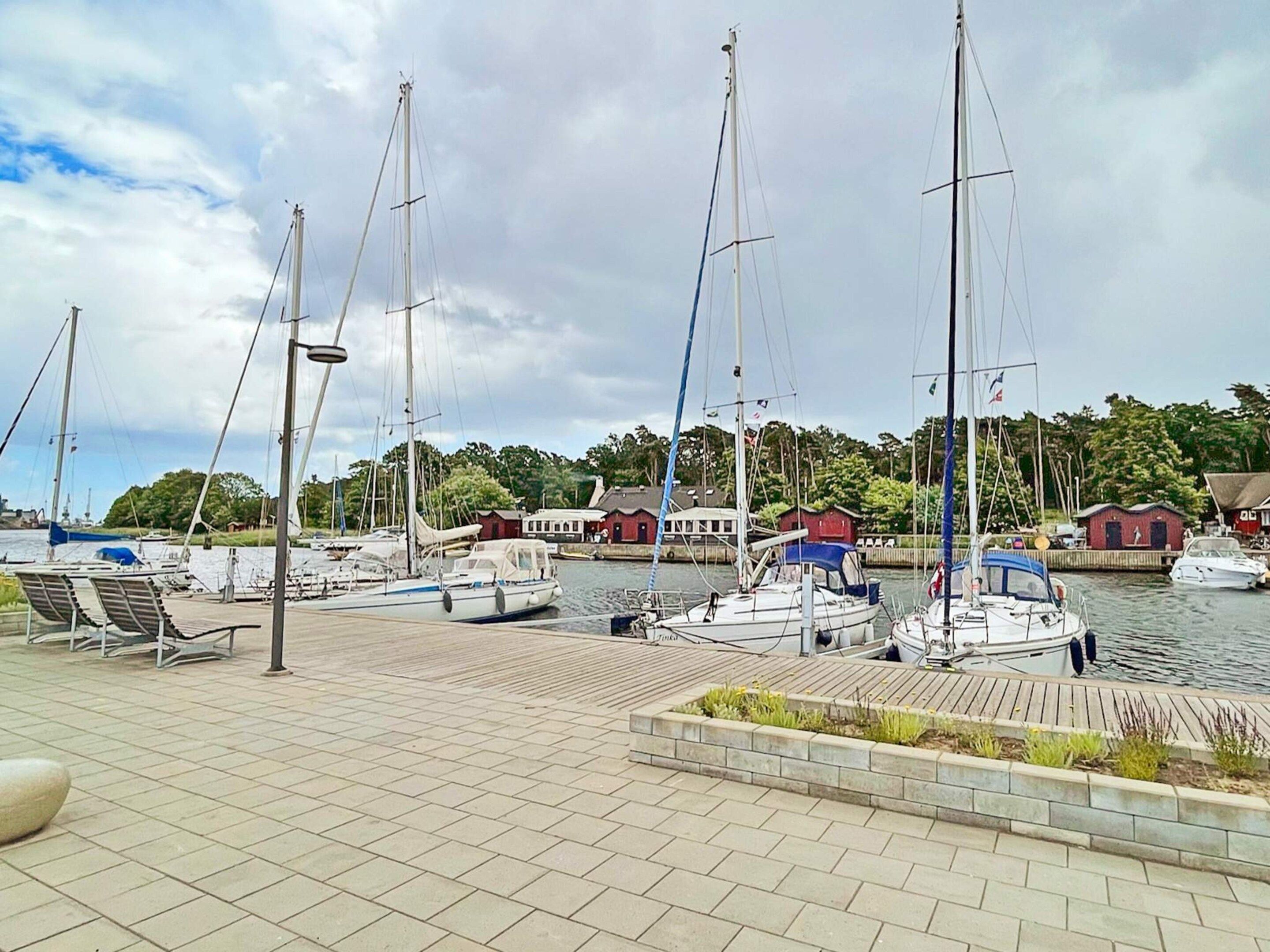 Un puerto deportivo con varios veleros amarrados, un muelle de madera y edificios al fondo.