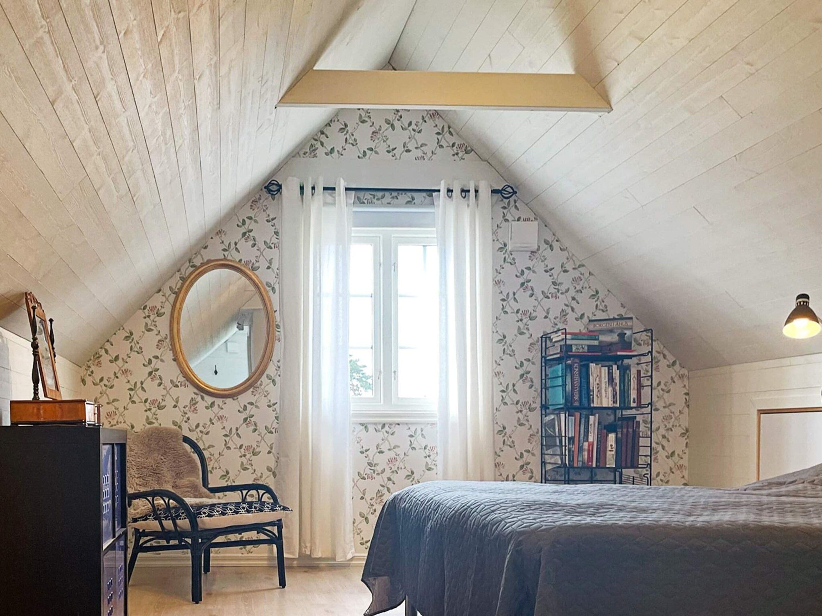 Un dormitorio con techo inclinado, una cama, una silla, una estantería, un espejo y una ventana con cortinas.