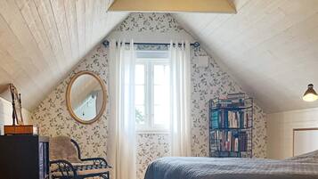 Un dormitorio con techo inclinado, una cama, una silla, una estantería, un espejo y una ventana con cortinas.
