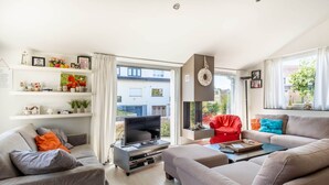 Ein modernes Wohnzimmer mit Flachbildfernseher, grauen Sofas und einem roten Sessel. Regale mit Dekorationselementen und ein Fenster mit Blick auf Gebäude.