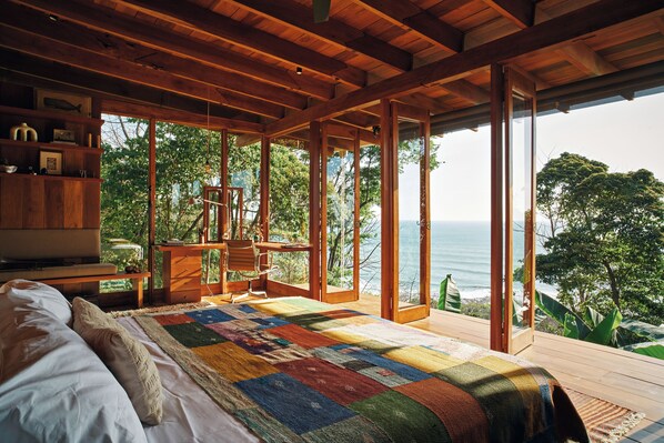Un dormitorio con una cama grande, vigas de madera, vista al océano y un escritorio de madera con una silla.