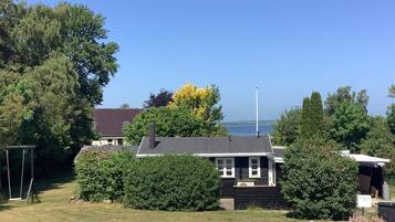 A house with a dark roof, white trim, and a chimney. It is surrounded by greenery and a body of water is visible in the background.