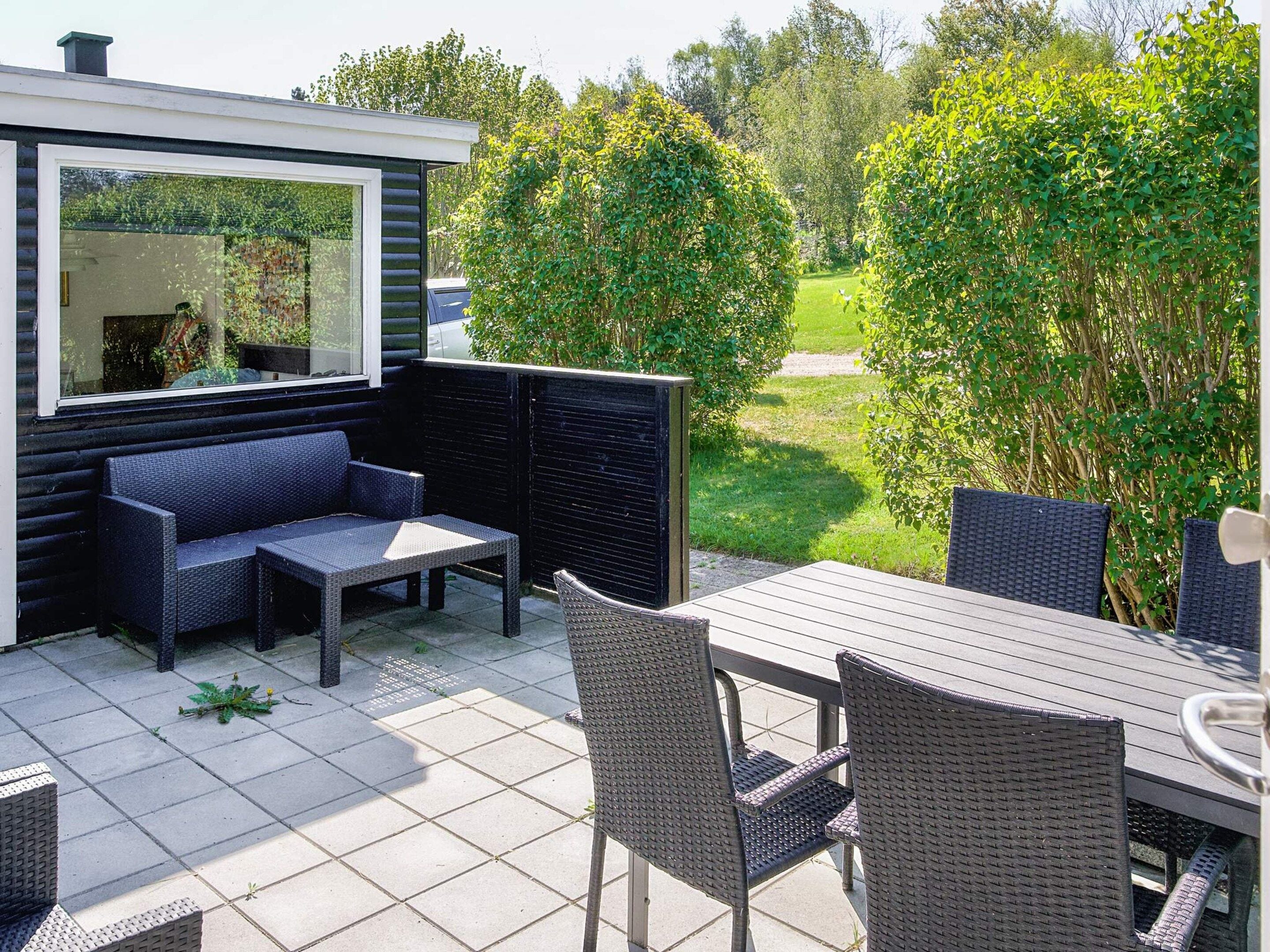 A patio with a sofa, table, and chairs, surrounded by a garden and a hedge.