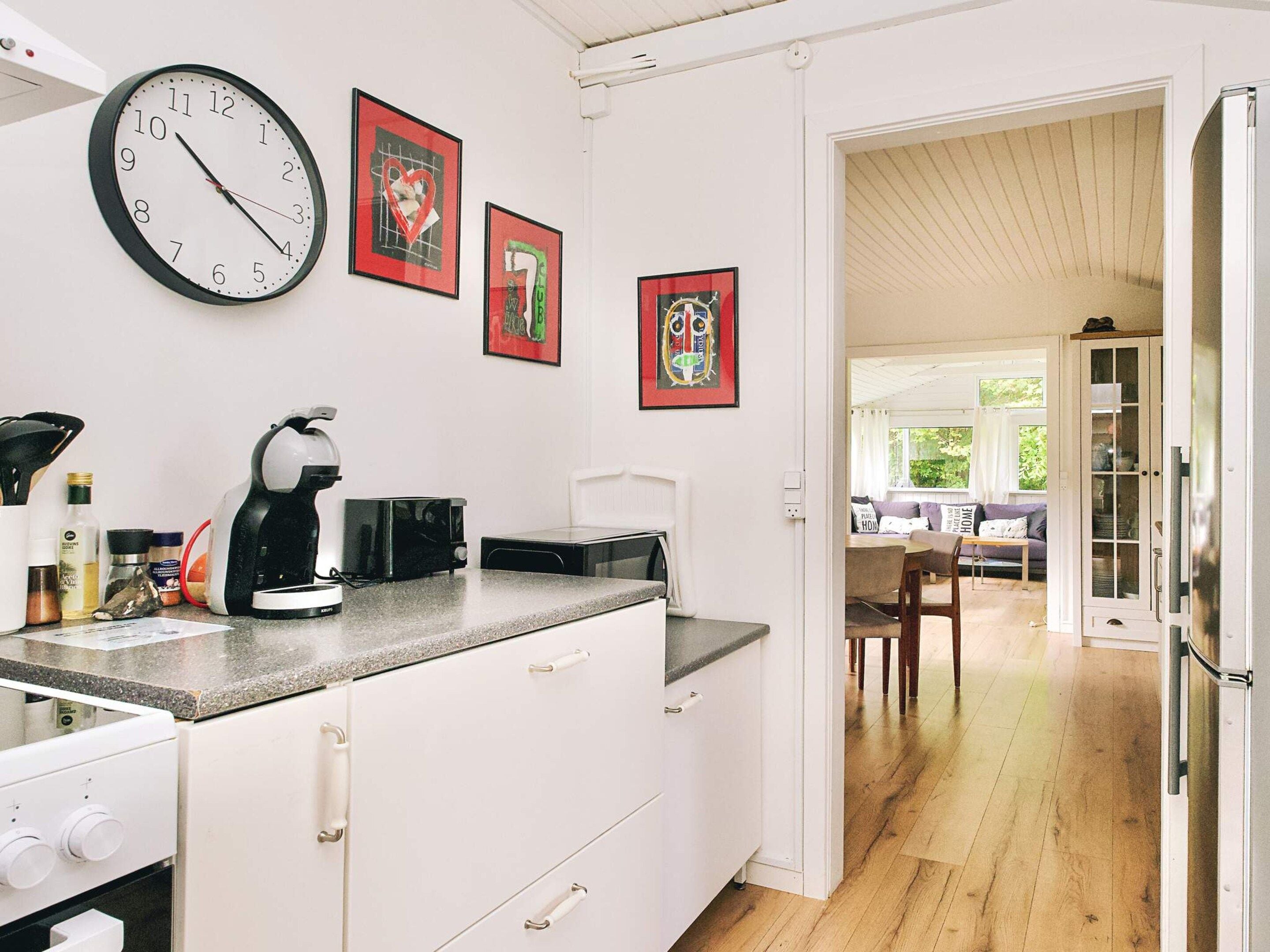 Una cocina con un reloj, microondas, hervidor y armarios. Hay cuadros en la pared y una vista hacia un comedor.