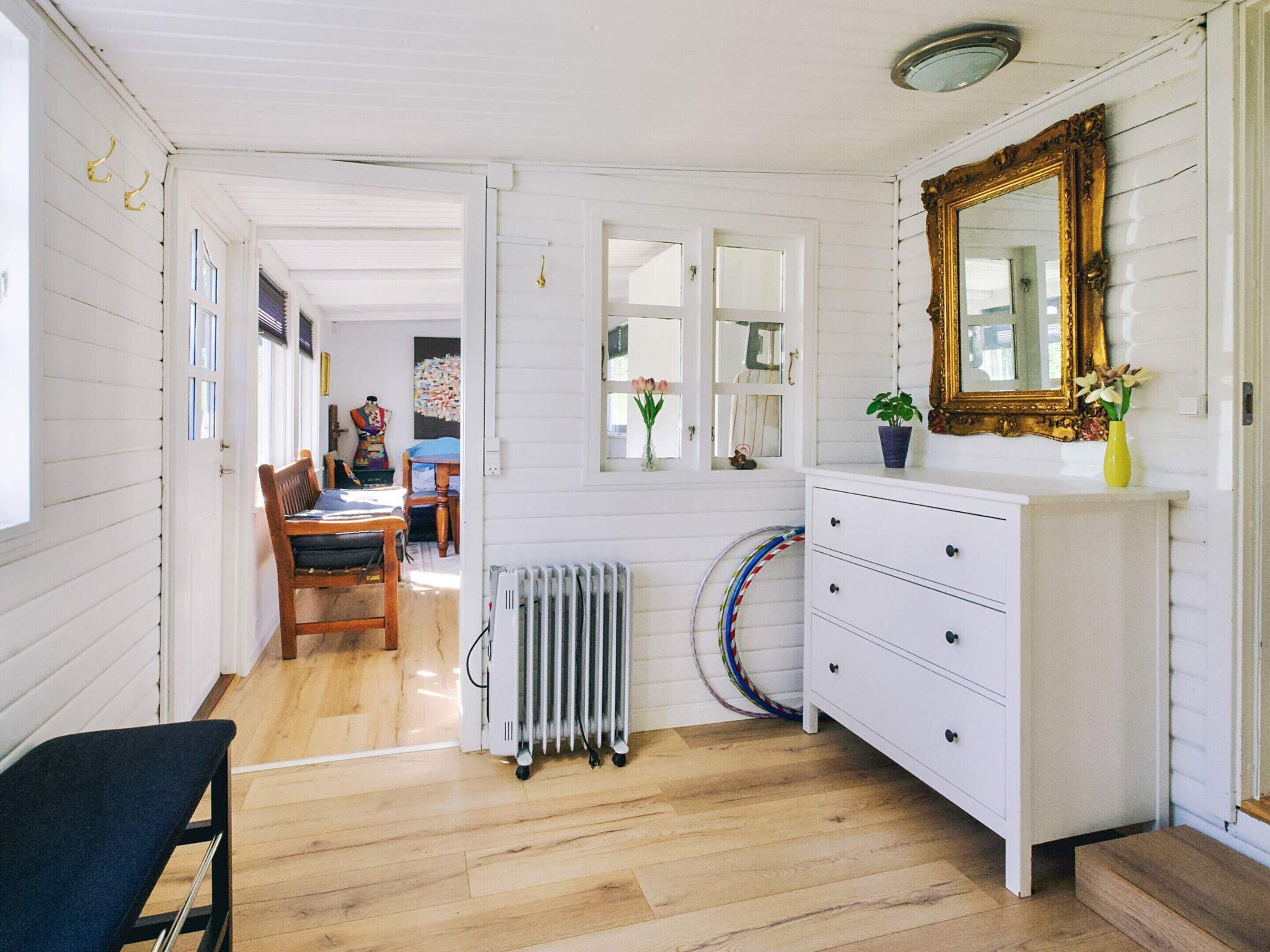 Una habitación con piso de madera, paredes blancas, un radiador, un tocador blanco, un espejo y una puerta que da a otra habitación.