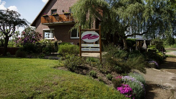 A two-story brick house with a balcony, surrounded by lush greenery and flowering plants. - Heidetraum in der Pension Heidetraum (Bispingen)