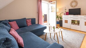 A modern living room with a blue sofa, a small white table, and a kitchen area with a TV and cabinets. - Heidetraum in der Pension Heidetraum (Bispingen)