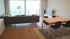 Moderni olohuone, jossa on sohva, ruokapöytä tuoleineen, televisio ja suuri ikkuna, jossa on verhot.