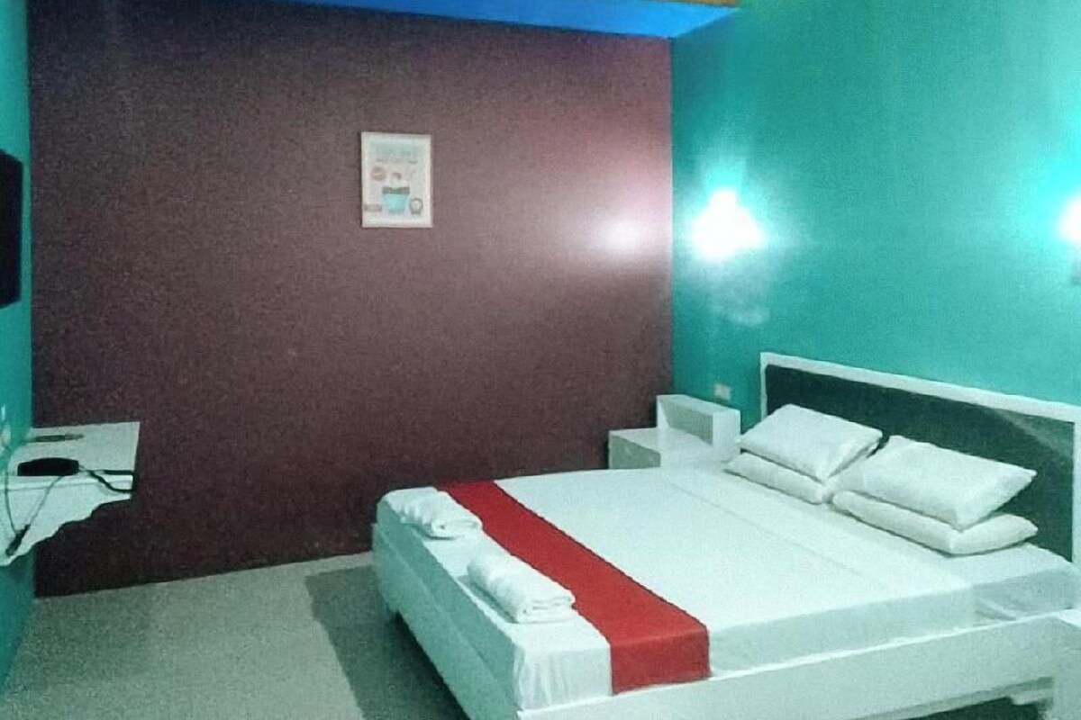 Un lit simple avec une literie blanche et un élément décoratif rouge, une table de chevet et un luminaire fixé au mur.