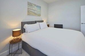 A modern bedroom with a large bed, a side table with a lamp, a radiator, and a painting on the wall.