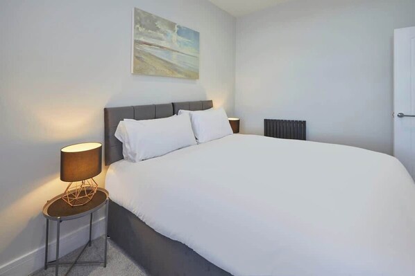 A modern bedroom with a large bed, a side table with a lamp, a radiator, and a painting on the wall.