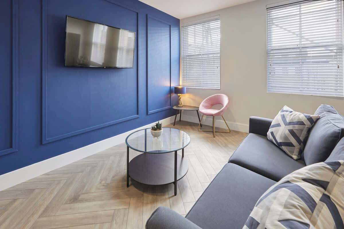 A modern living room with a blue accent wall, a round glass coffee table, a grey sofa, and a flat-screen TV mounted on the wall.