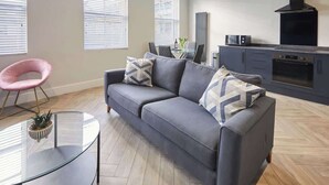 A modern living room with a grey sofa, a glass coffee table, and a pink chair. There is a kitchen area with a stove and oven. - Anya’s Lookout - 1 Bedroom Apartment In Scarbourgh (North Yorkshire)