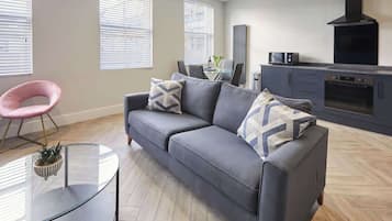 Un salon moderne avec un canapé gris, une table basse en verre et un fauteuil rose. Il y a également un coin cuisine avec une cuisinière et un four.
