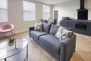 En moderne stue med en grå sofa, et glasbord og en lyserød stol. Der er et køkkenområde med komfur og ovn.