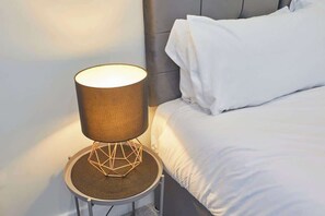 A modern bedside table with a lamp, a bed with white bedding, and a grey headboard.