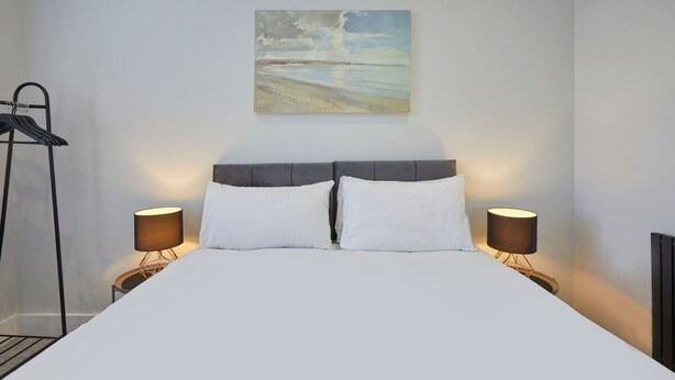 A neatly made bed with white linens, two bedside lamps, and a painting above the headboard. - Anya’s Lookout - 1 Bedroom Apartment In Scarbourgh (North Yorkshire)