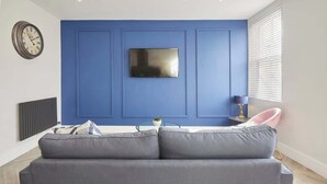 A living room with a blue wall, a flat-screen TV mounted above a grey sofa, a round clock on the wall, and a window with blinds. - Anya’s Lookout - 1 Bedroom Apartment In Scarbourgh (North Yorkshire)
