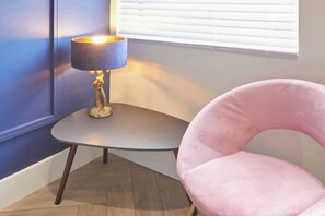 A corner room with a small round table, a pink chair, and a lamp.