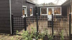 A small, dark-colored building with two windows, a fenced area with plants, and a tree in the background. - 4 Star Holiday Home in Norre Nebel (Norre Nebel)