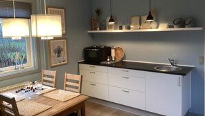 A compact kitchen with white cabinets, a black microwave, and a sink. There are two pendant lights, a window with blinds, and a dining table with chairs. - 4 Star Holiday Home in Norre Nebel (Norre Nebel)