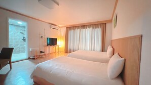 Habitación de hotel con dos camas, un televisor, un escritorio y un baño visible a través de una puerta de cristal.