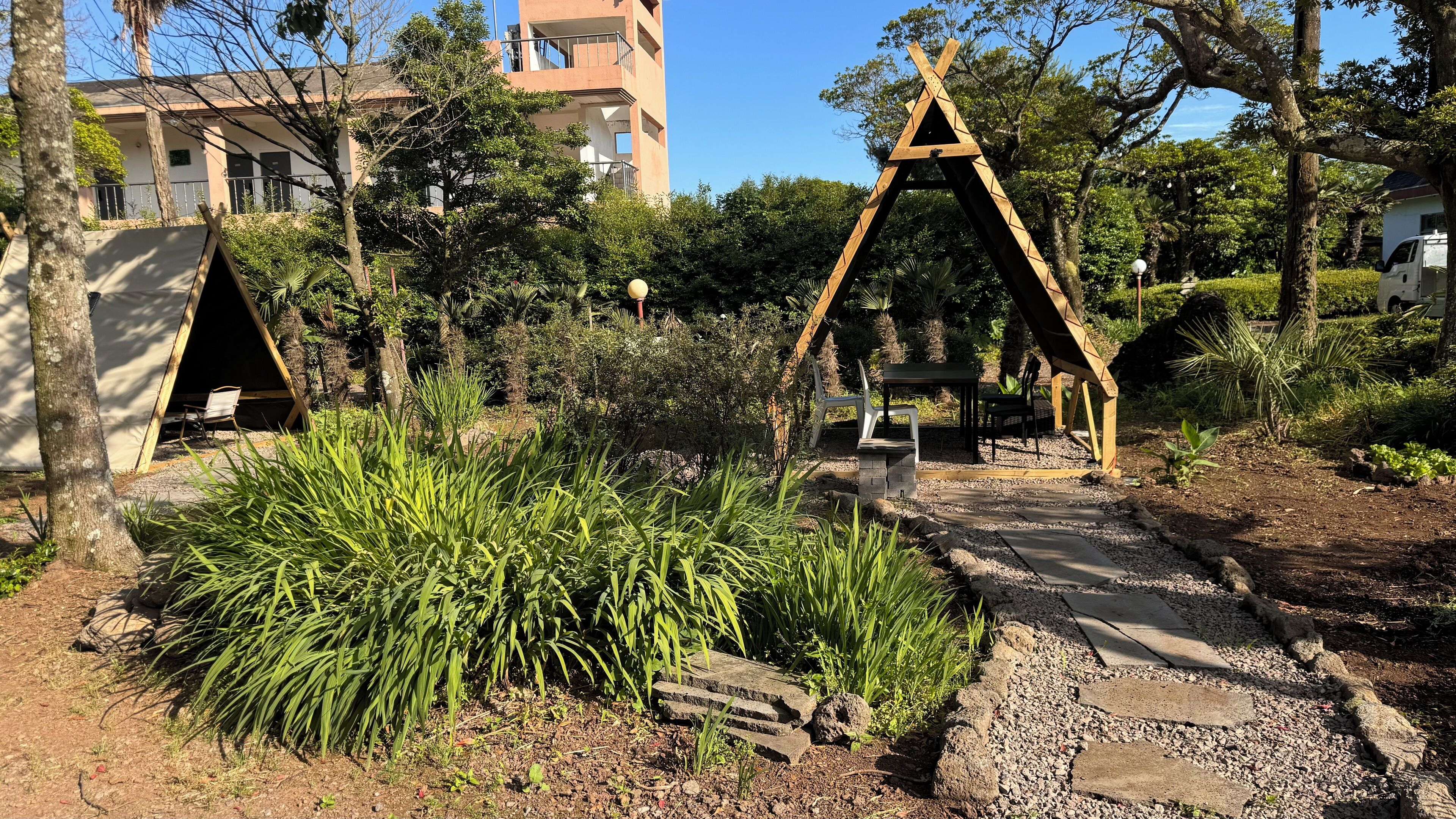 A garden with a wooden swing set, a small table, and chairs. There are trees and a building in the background.