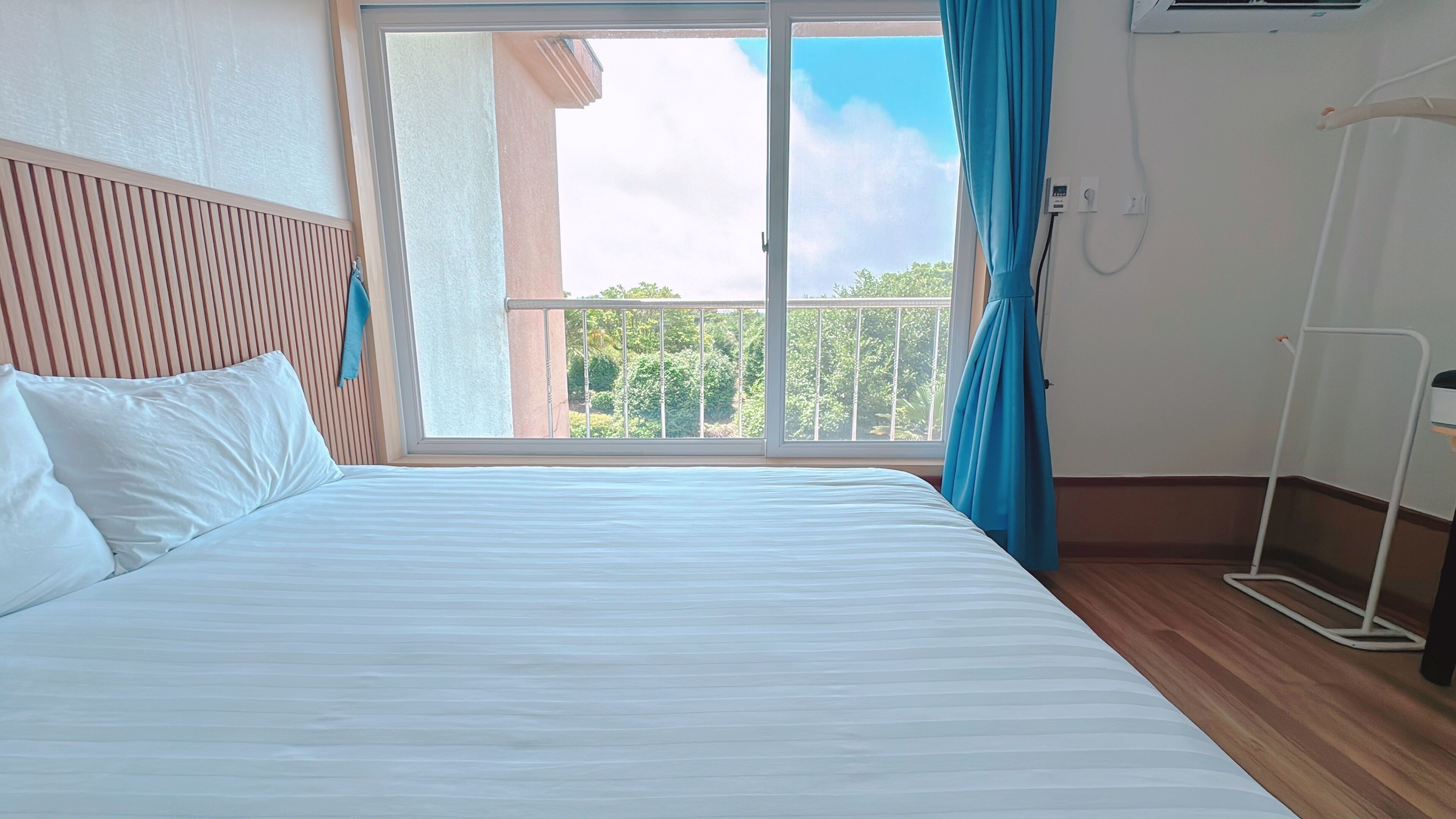 A hotel room with a bed, a window with a view of greenery, and an air conditioning unit.