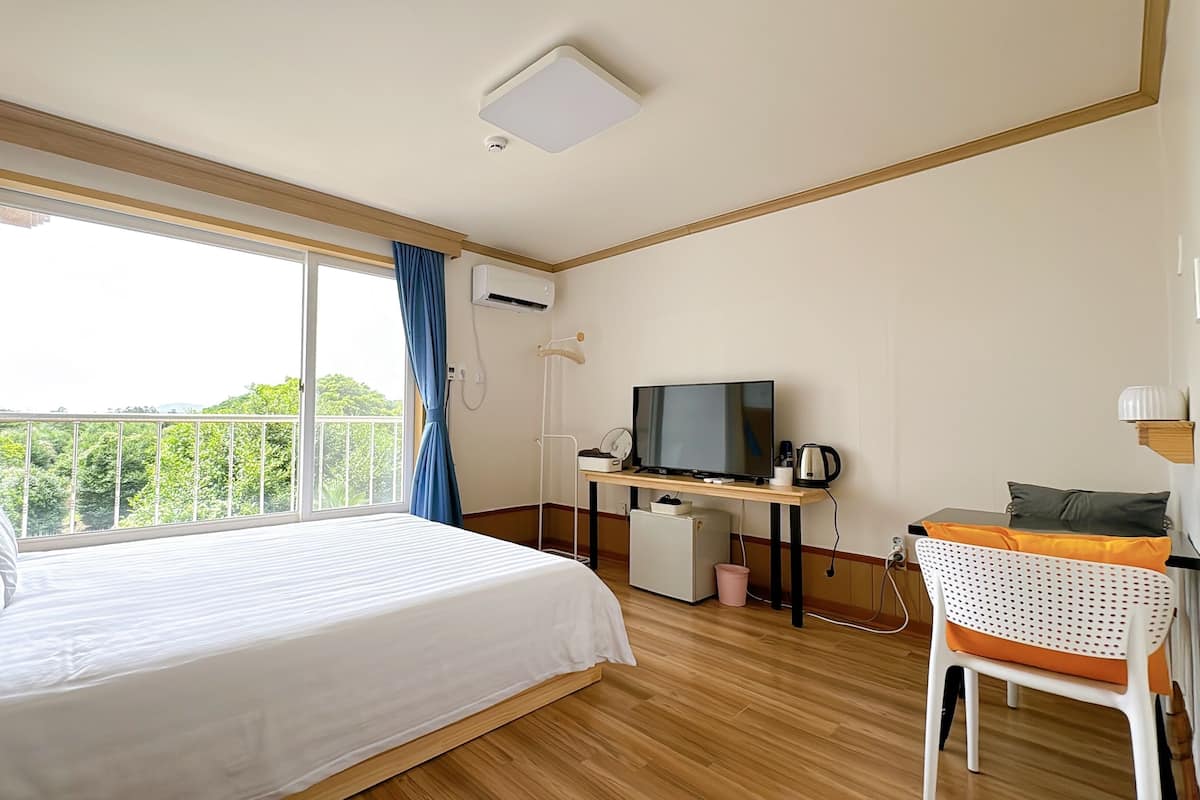 A hotel room with a bed, a desk with a television, a chair, and a view of greenery outside.