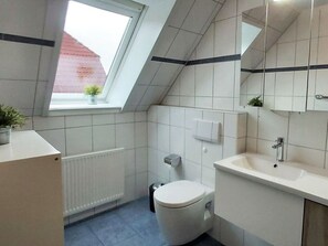 A modern bathroom with a skylight, white tiles, a toilet, a sink, and a mirror. - Holiday Apartment East Dunes (Borkum)