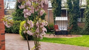 Un chemin pavé de briques mène à un espace patio agrémenté d’un petit arbre et d’une table avec des chaises.