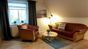 Un salon avec un canapé en cuir marron, un fauteuil en cuir marron, une petite table basse ronde et un lampadaire. Il y a une fenêtre avec des rideaux et un radiateur en dessous.