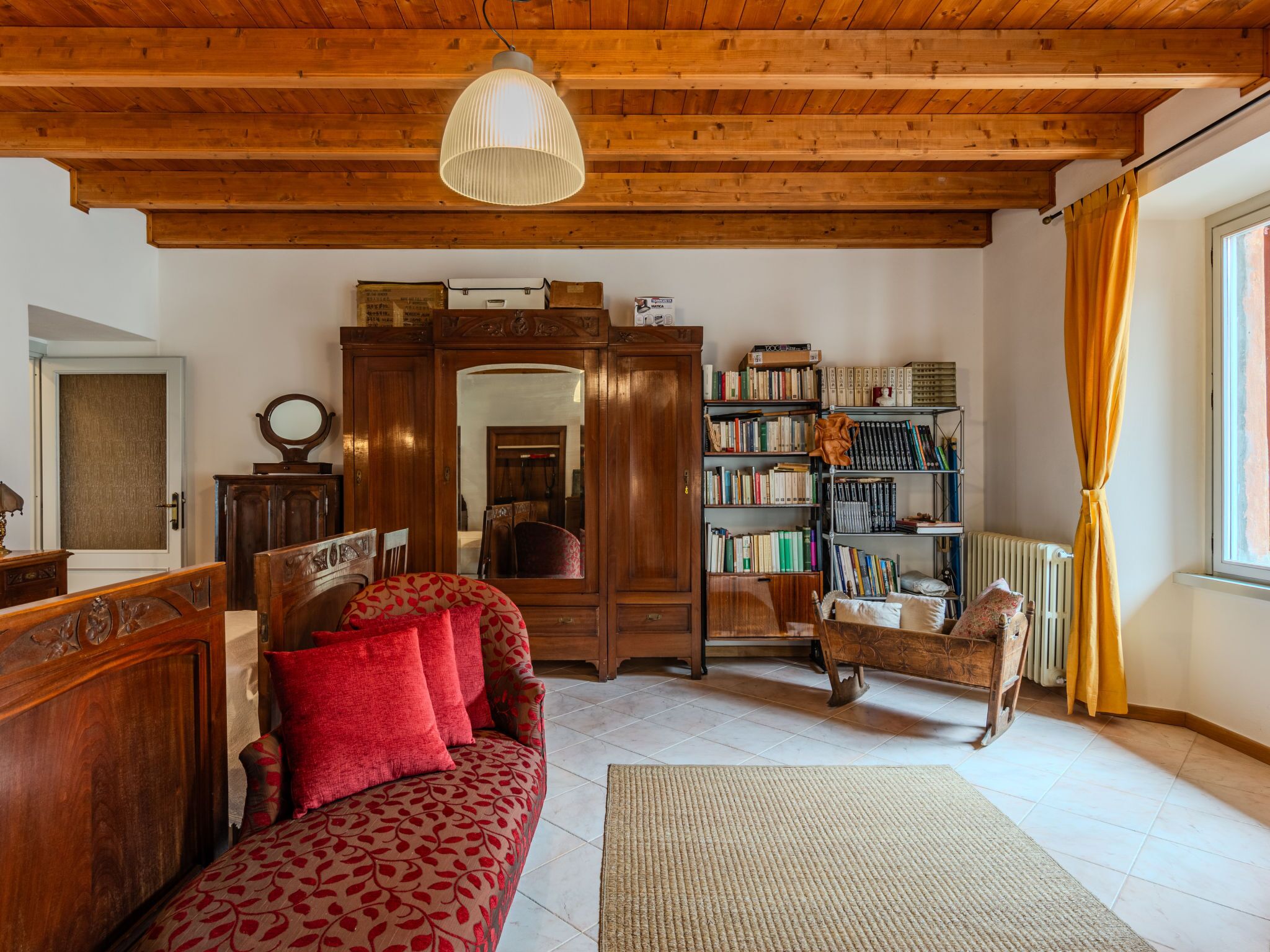 A cozy room with a wooden ceiling, a sofa with red cushions, a bookshelf, and a radiator.