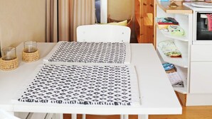 A small, neatly organized room with a white table, two patterned placemats, a white chair, a shelf with various items, and a window with curtains. - 4 Person Holiday Home in Falkenberg-by Traum (Falkenberg)