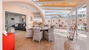 A spacious living area with a glass dining table surrounded by wicker chairs, a red cabinet, and a stone planter with flowers.